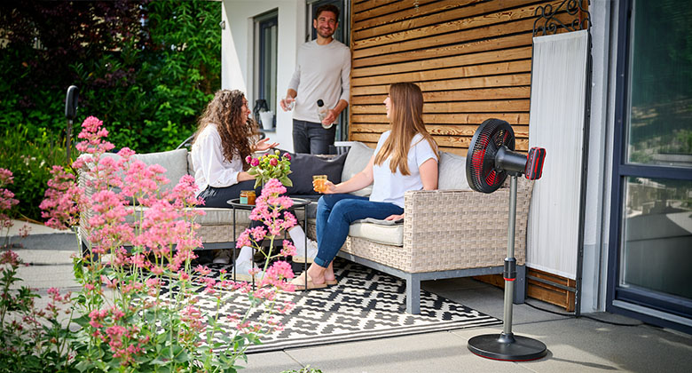 Drei Personen sitzen auf einer Terrasse neben einem schwarzen Standventilator mit rotem Schutzgitter und trinken Getränke.