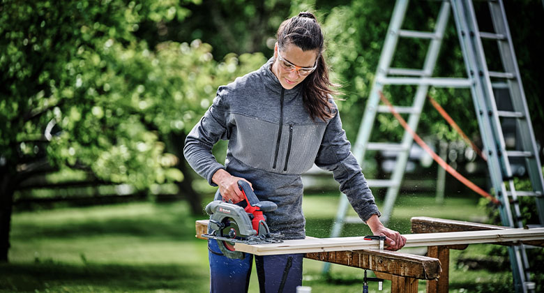 Frau mit Schutzbrille sägt im Garten ein Holzbrett mit einer roten Handkreissäge auf zwei Böcken.