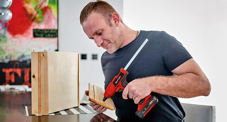 Mann trägt mit einer roten Akku-Klebepistole Klebstoff auf ein Holzbrett auf und hält ein weiteres Brett in der Hand.