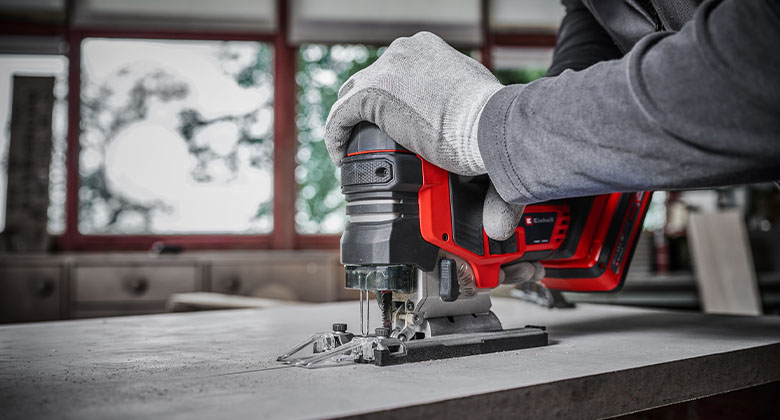 Hand mit Arbeitshandschuh führt eine rote Akku-Stichsäge durch eine Holzplatte.