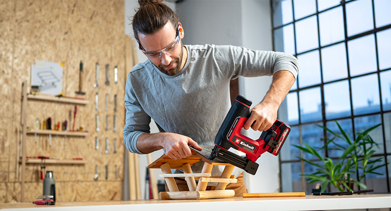 Mann mit Schutzbrille befestigt Holzteile mit einem roten Akku-Nagler auf einer Werkbank.